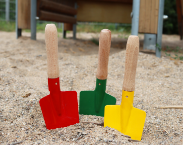 Preview: Drei Sandschaufeln für Kinder stecken nebeneinander im Sand eines Spielplatzes. Sie haben einen roten, einen grünen und einen gelben Besatz.